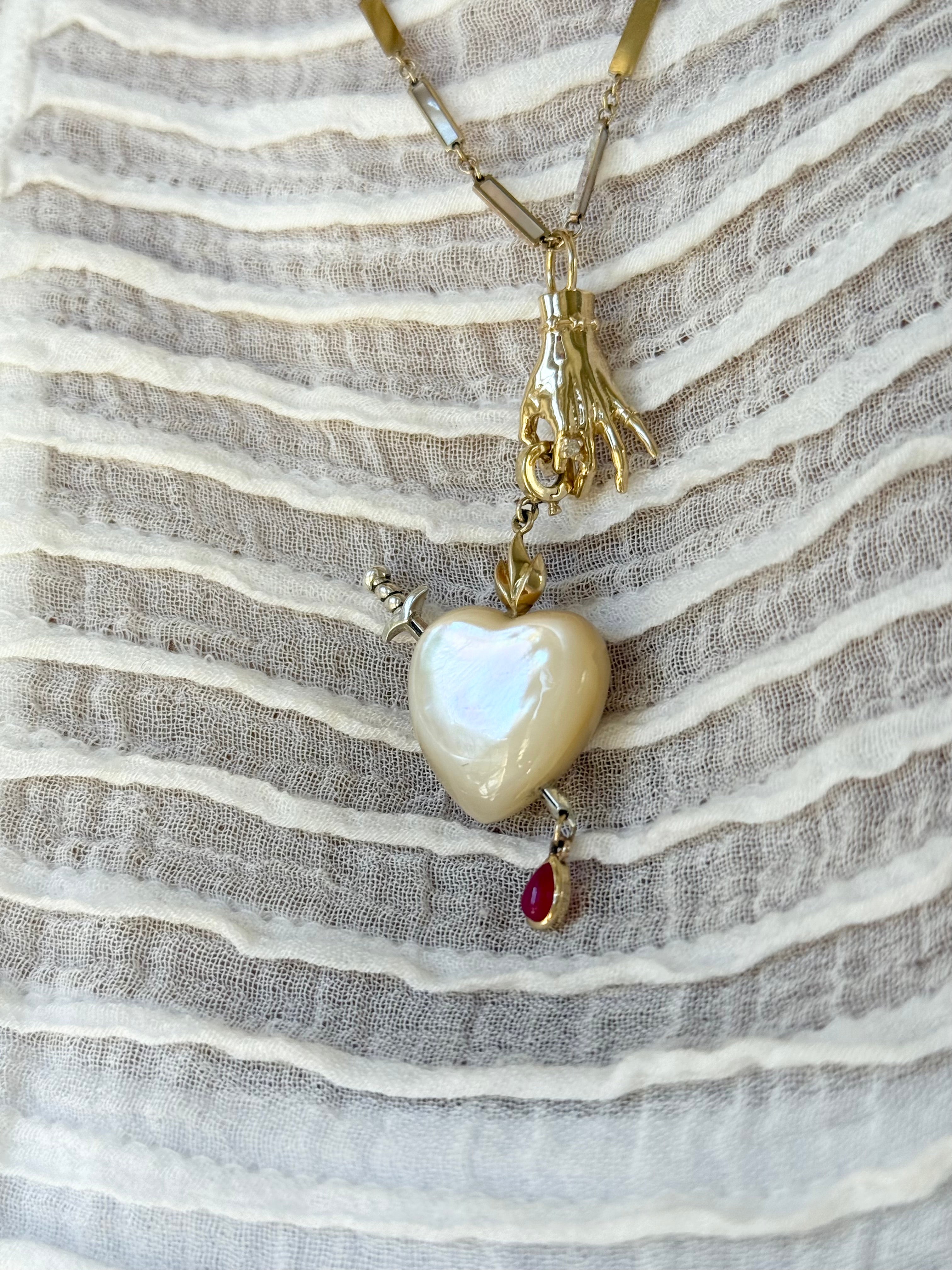 Maura Green Jewelry gyan mudra ladyfinger charm dangling on a gold chain. It's fingers are holding a white mother of pearl sacred heart charm featuring a silver sword and gold flames. A teardrop shaped ruby dangles from the tip of the sword.