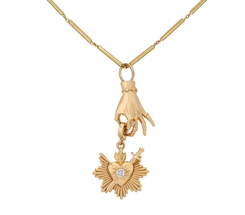 A gold pendant necklace featuring a hand holding a sacred heart pendant with a diamond at the center, displayed on a clean white background. Designed by Maura Green Jewelry