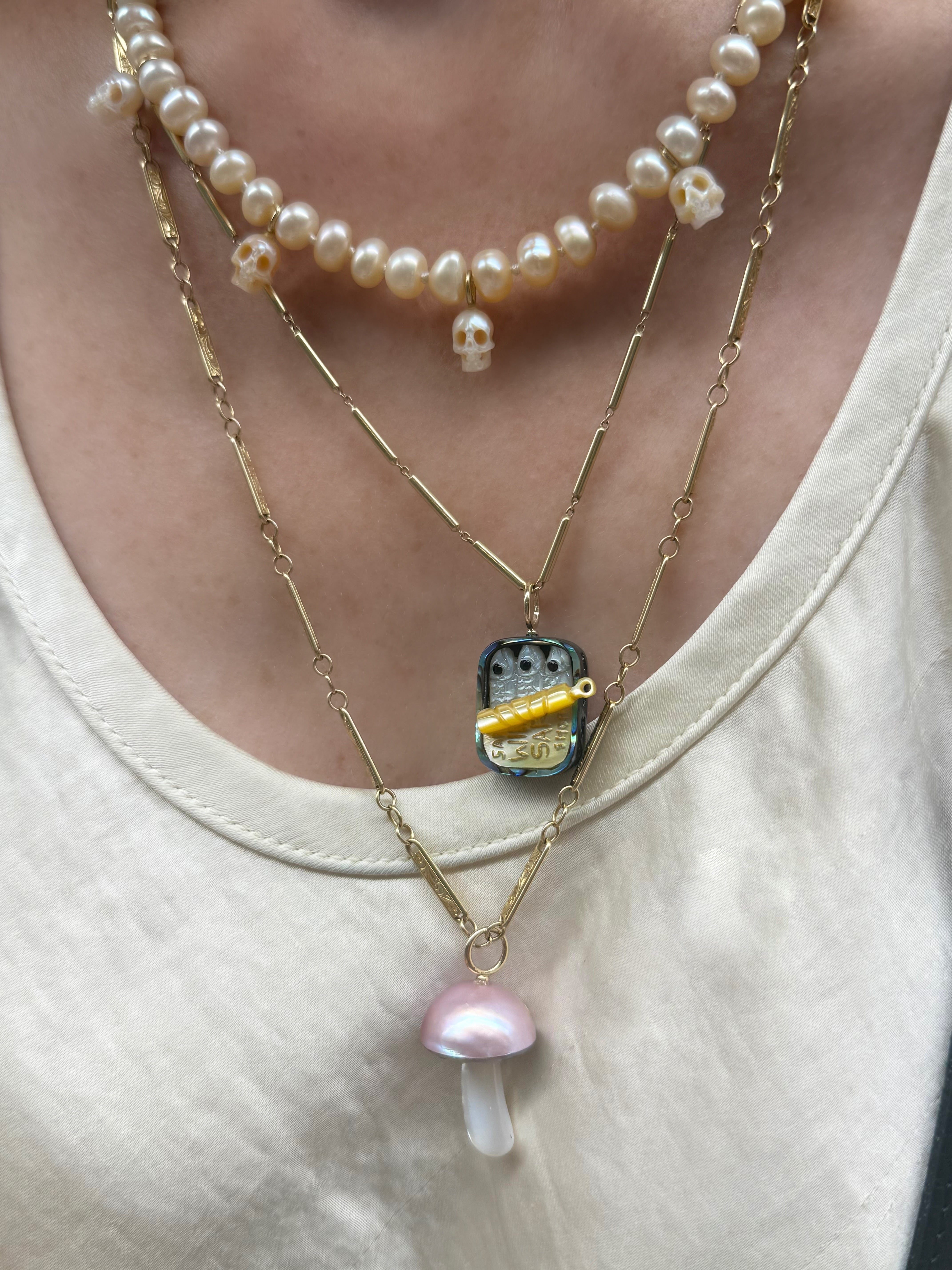 Close-up of a Maura Green wearing two necklaces featuring a sardines charm, a pink pearl mushroom charm, and a strand of pearls with skulls, with a white shirt background