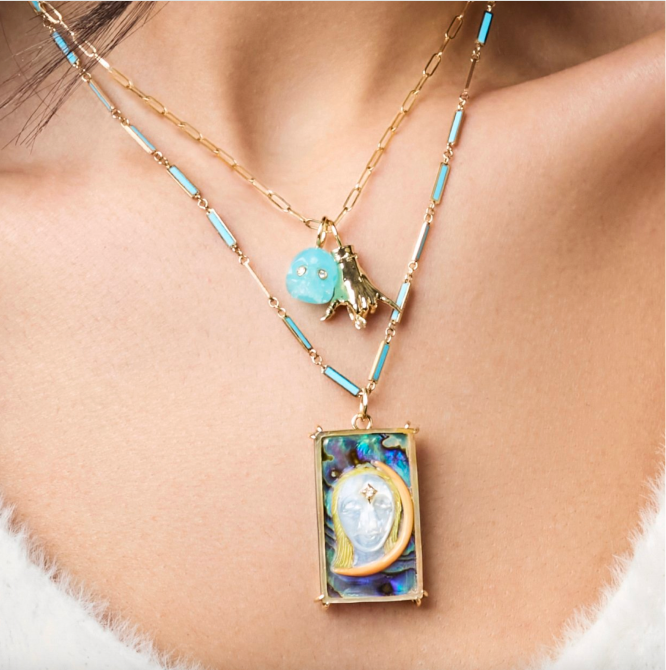 Cropped shot of layered necklaces on a woman's chest, with various charms designed by Maura Green Jewelry, including a shaka ladyfinger, a high priestess tarot card charm, and a blue skull charm.