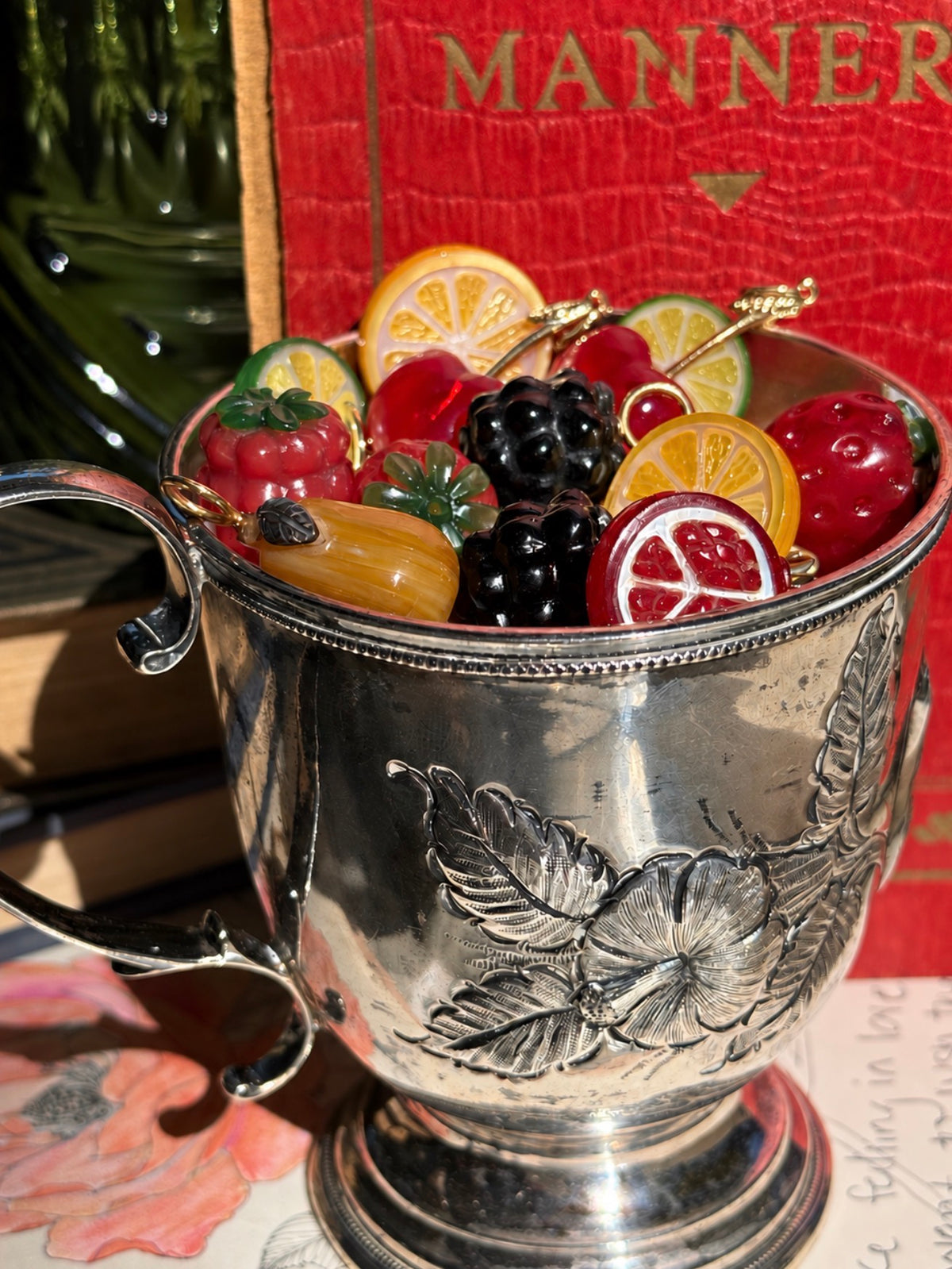 An antique sterling silver cup with.a scrollwork handle and repousse engraved flowers is tilted to show that it is filled with colorful hand carved fruit and berry charms designed by Maura Green. The charms include  golden mother of pearl lemons, red chalcedony cherries with golden stems, golden mother of pearl pears, a red pomegranate, as well as blackberries and raspberries.  The background there is a red antique book with a leather cover which says the word "Manners" in embossed in gold.