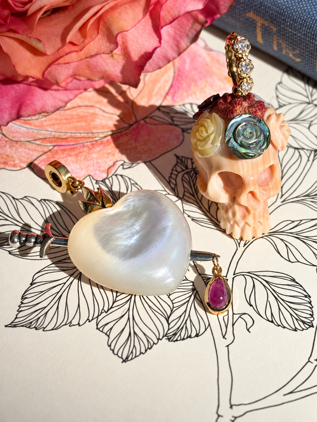Two charm are laid upon a book with a black and white illustration of leaves. In the background is a wilting pink rose and the spine of a blue antique book. One charm is a hand carved apricot conch skull crowned with colorful carved flowers, with a gold floral bail with diamonds. The second charm is a white mother of pearl sacred heart, which is pierced by a silver sword with a teardrop shaped ruby dangling from it's tip. Designed by Maura Green.