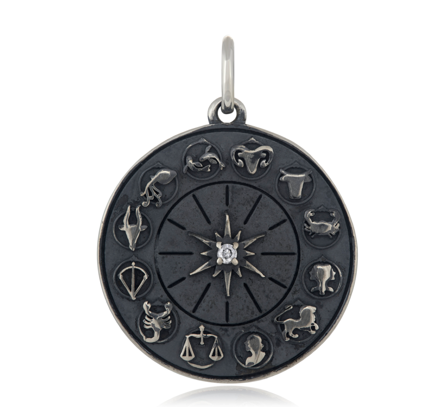 An oxidized sterling silver zodiac wheel charm with a prong set white diamond in the raised starburst shape at it's center, featuring various zodiac signs around the circumference. The silver charm is in front of a white backdrop.