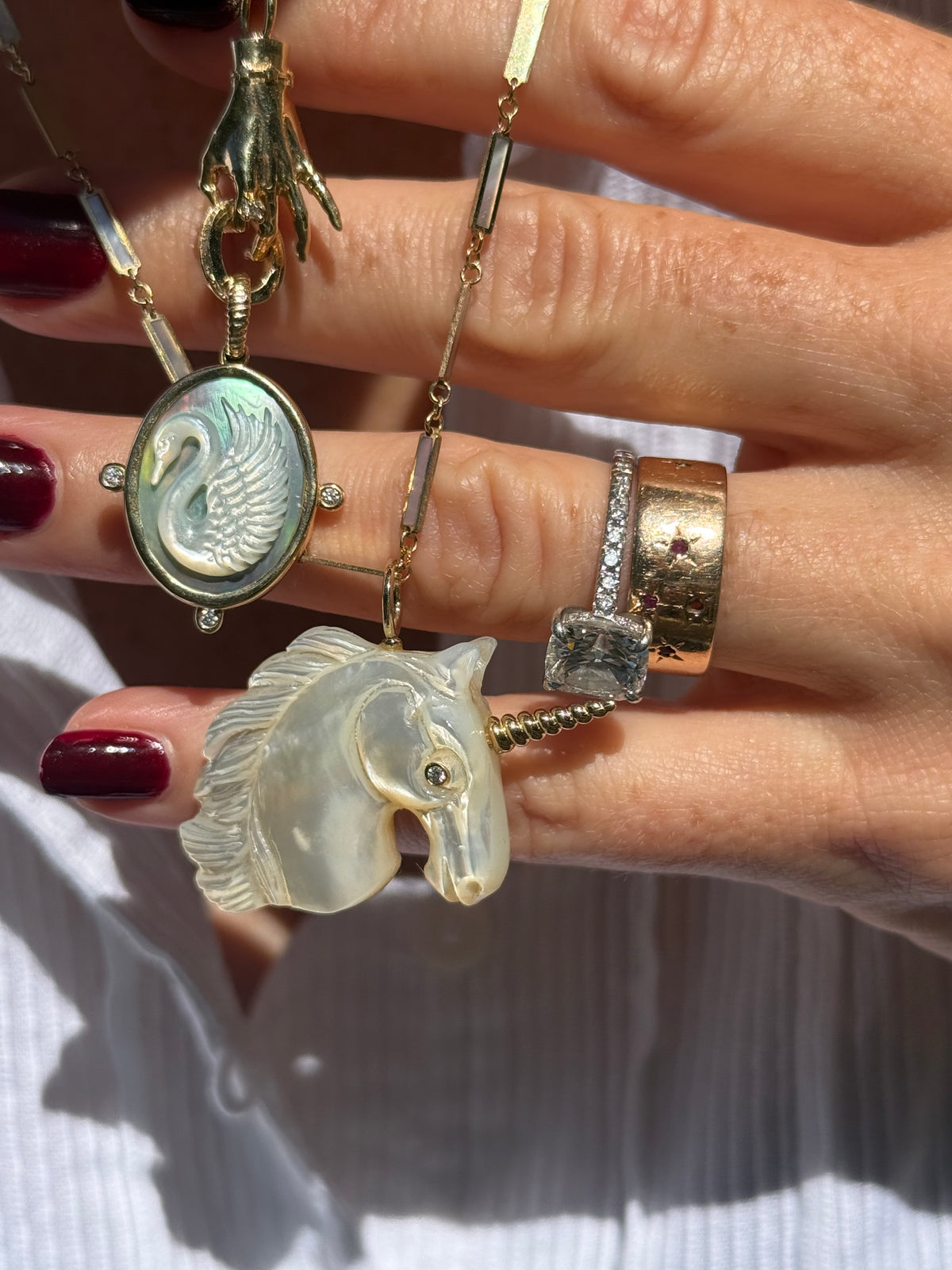 A close-up of a hand holding a gold-toned necklace with multiple charms: a round iridescent swan pendant, a translucent white horse head with a spiral horn, and a gold ring on the finger. Designed by Maura Green