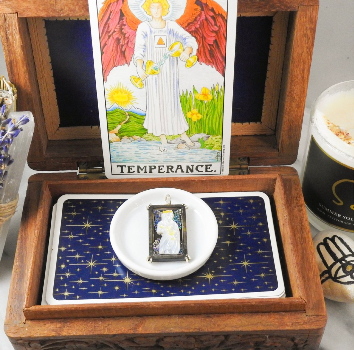 A rectangular charm depicting the temperance tarot card, sits atop a stack of paper tarot cards with a star pattern on them inside an open detailed carved wooden box.