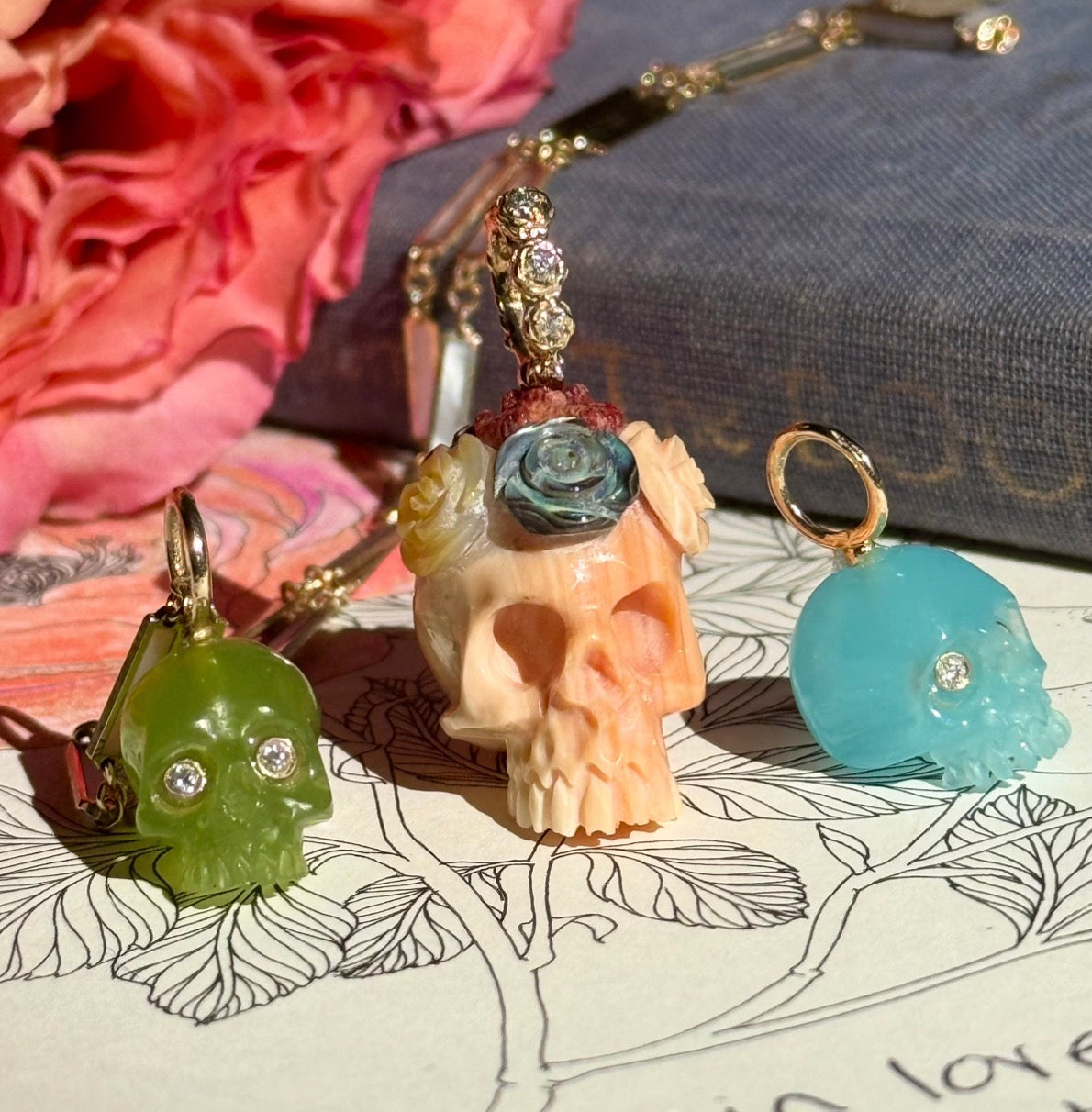 Three Hand carved skull charms rest on the page of a book with a pink rose in the background. The skulls are grass green chalcedony with diamond eyes, apricot conch shell with a crown of colorful carved flowers, and pacific blue chalcedony with diamond eyes.