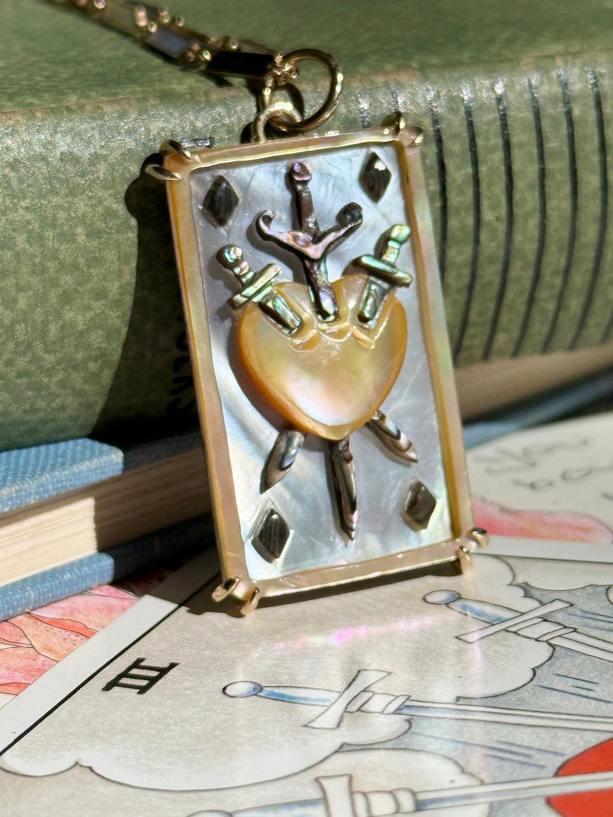 A rectangular hand carved tarot charm featuring a mother of pearl heart pierced by three abalone swords leans against the spine of a green book, and sits on top of a tarot card depicting the three of swords. The charm is held in the 4 corners by sterling siver prongs.