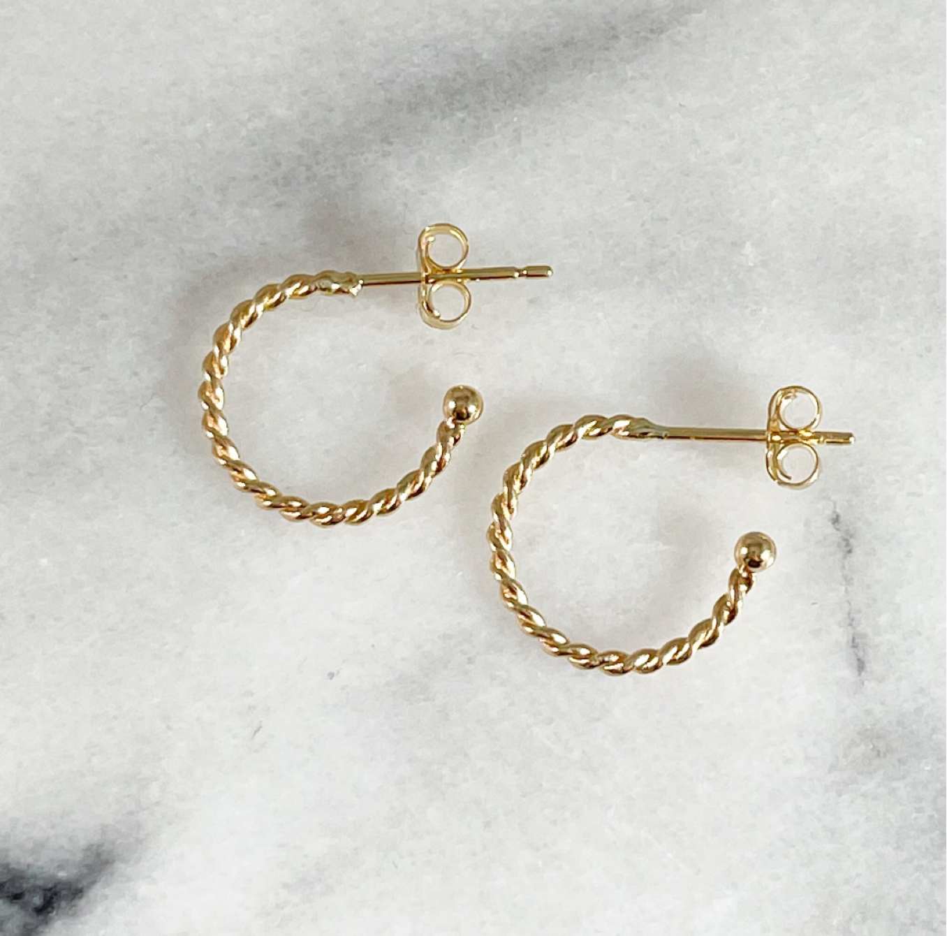 Side view of a pair of Maura Green Jewelry yellow gold hoop earrings with a twisted wire pattern and post and clutch closure against a white marble background.