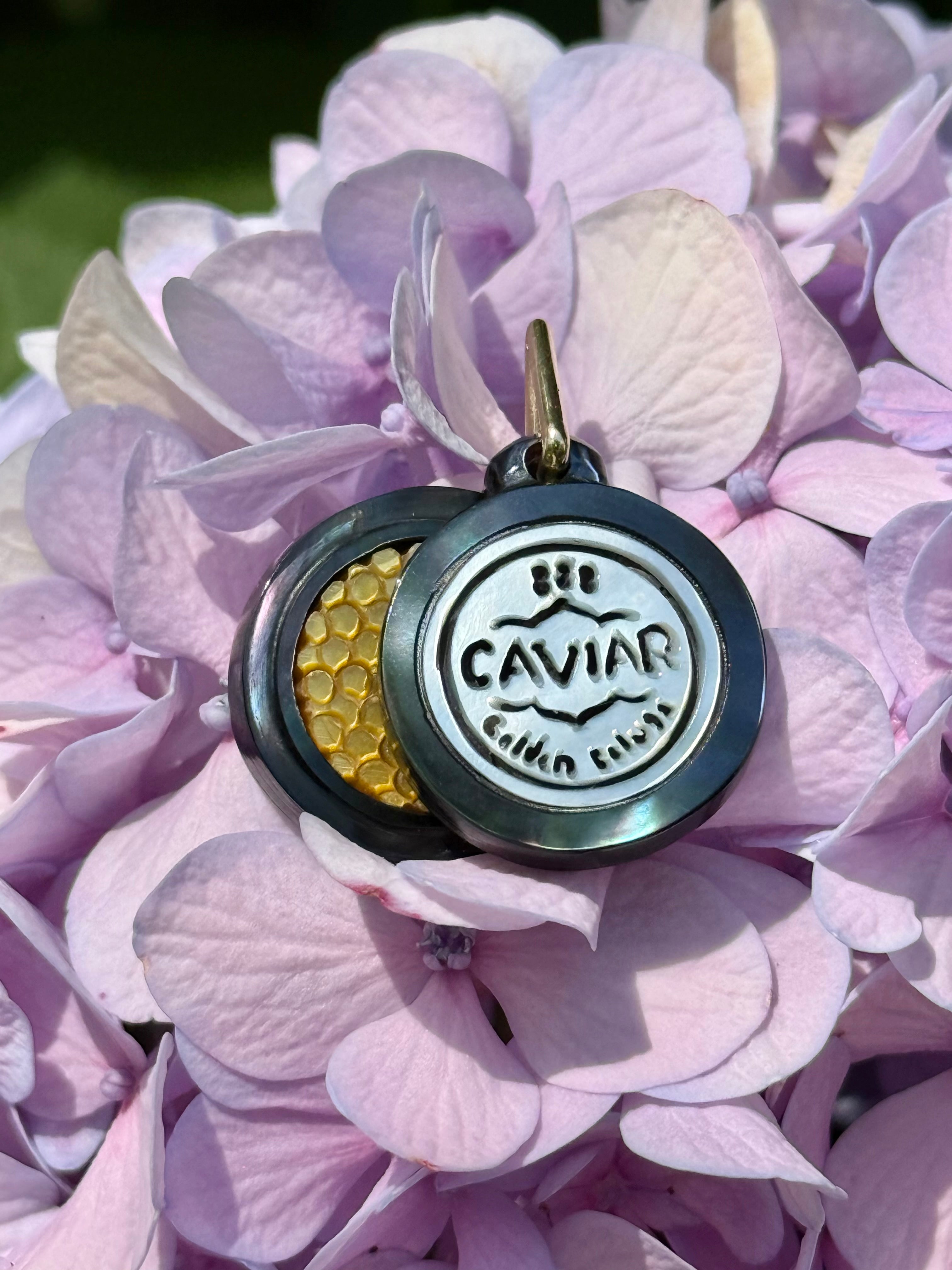 A round charm with a hinged lid, resting on pink and purple hydrangea petals. The front of the lid is silver-toned with the words 'CAVIAR' while the open side reveals a gold-toned honeycomb-textured interior. Designed by Maura Green Jewelry.