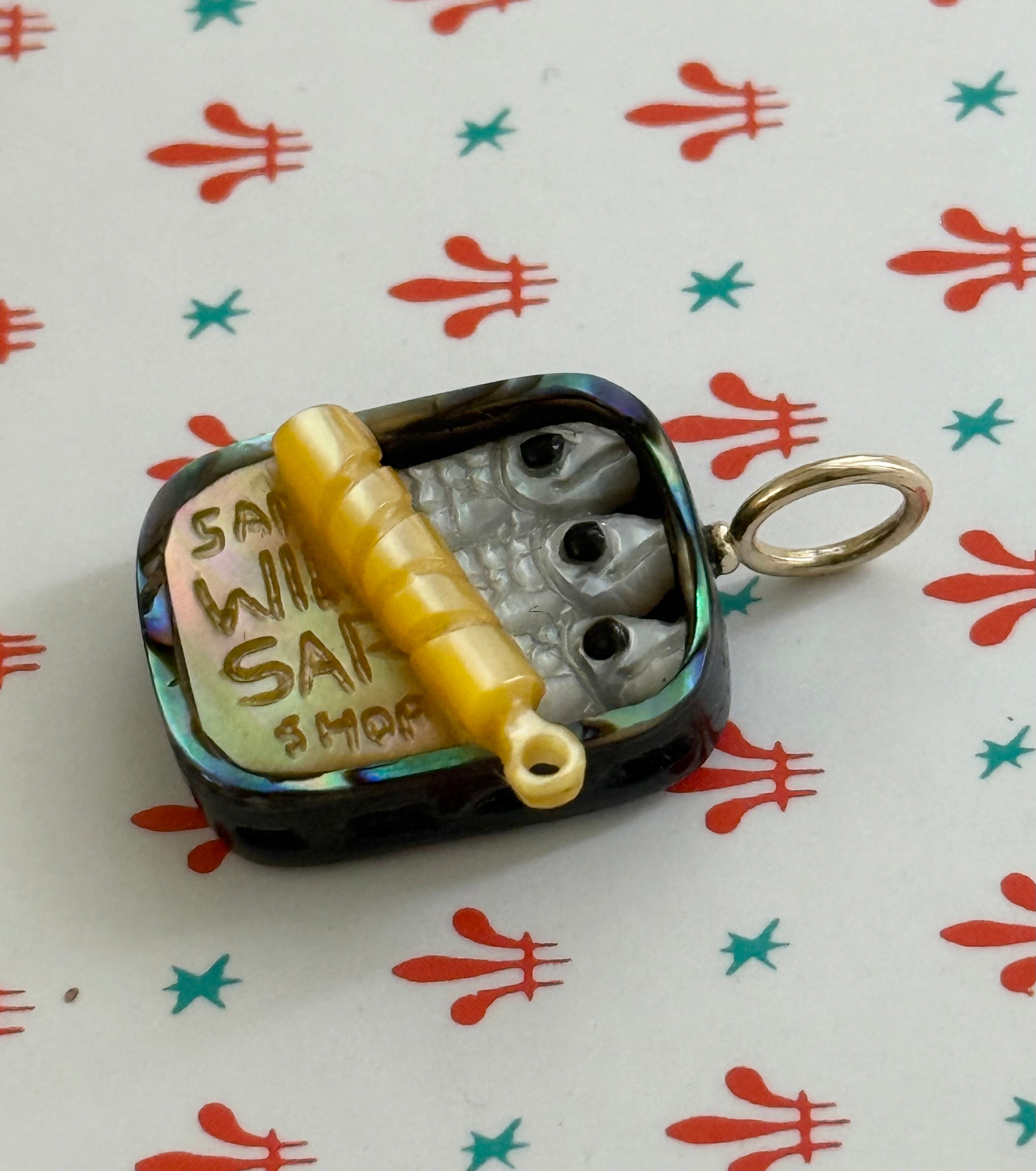 Small sardine tin charm featuring hand carved white mother of pearl fish in a colorful abalone tin against a patterned background.