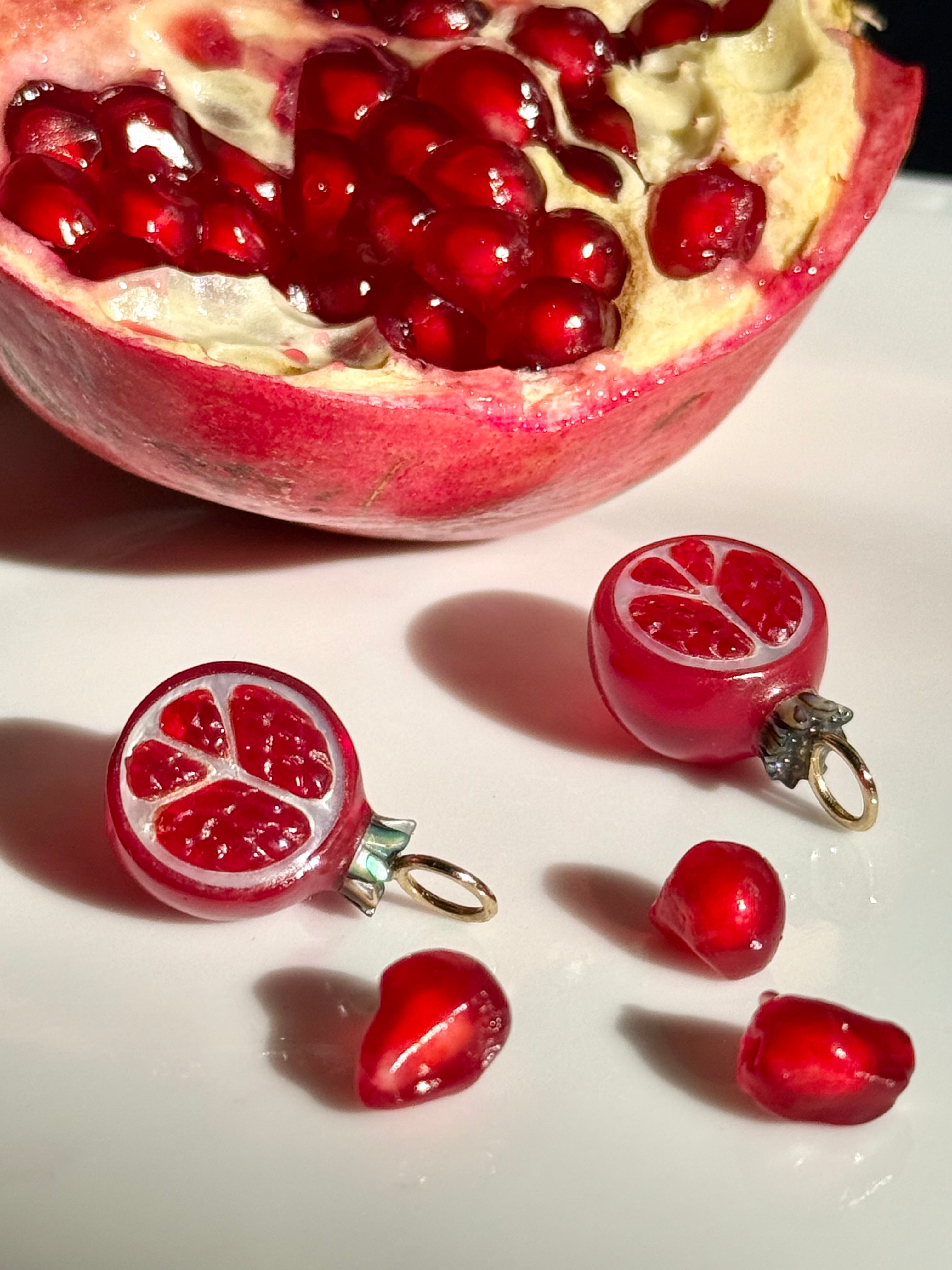 A halved pomegranate and red pomegranate-shaped jewelry charms by Maura Green on a white surface.