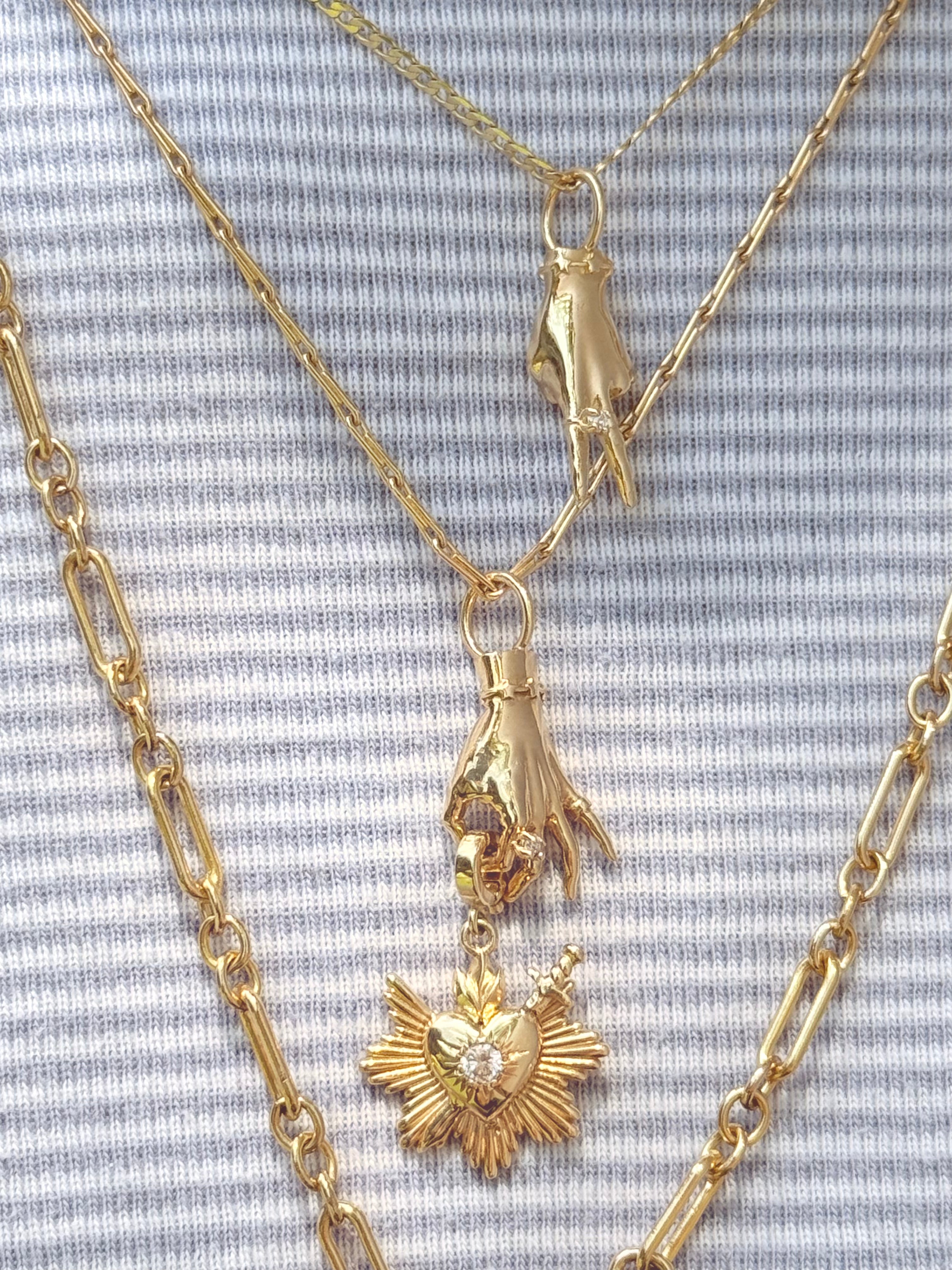 A layered gold necklace stack on a model wearing a light blue striped shirt. The set includes a delicate chain with a small hand-shaped pendant, a longer chain with a larger hand pendant holding a sacred heart charm with a diamond in the center. Designed by Maura Green Jewelry.