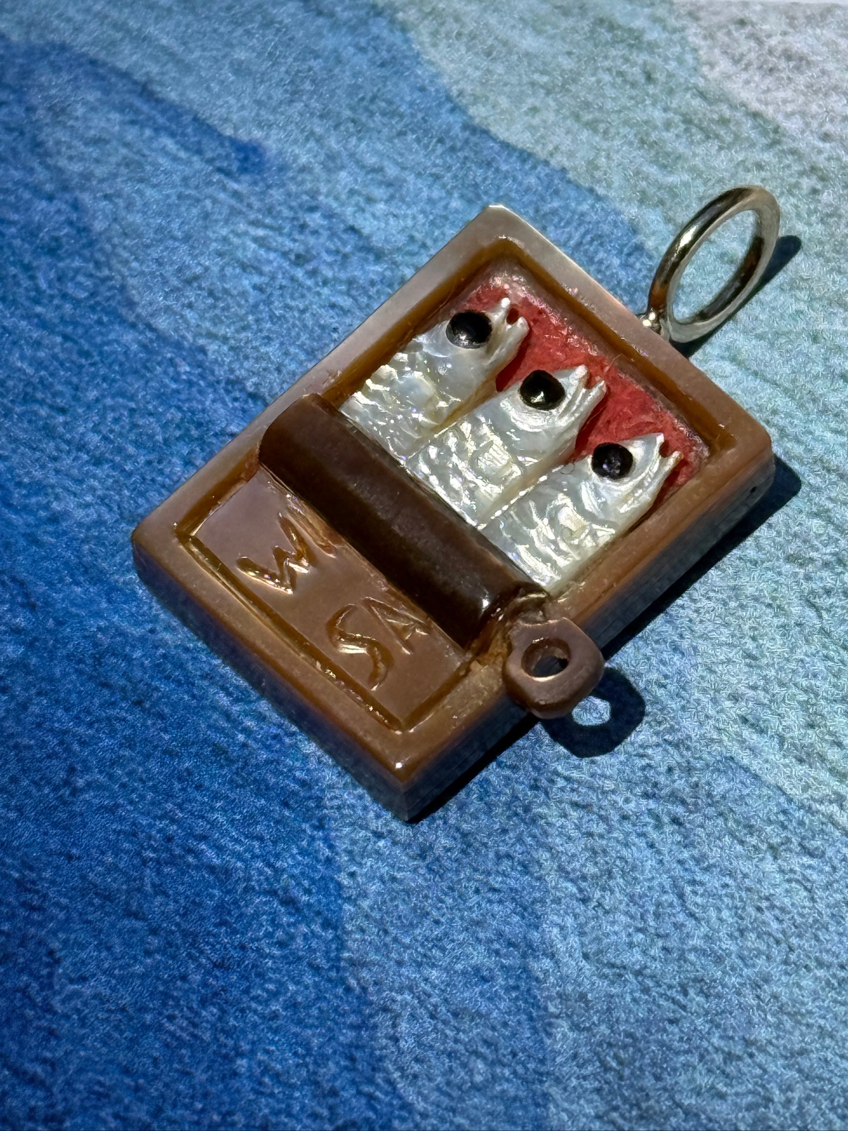 Brown tin with three white sardines on a 14k gold bail on a blue background. Designed by maura green