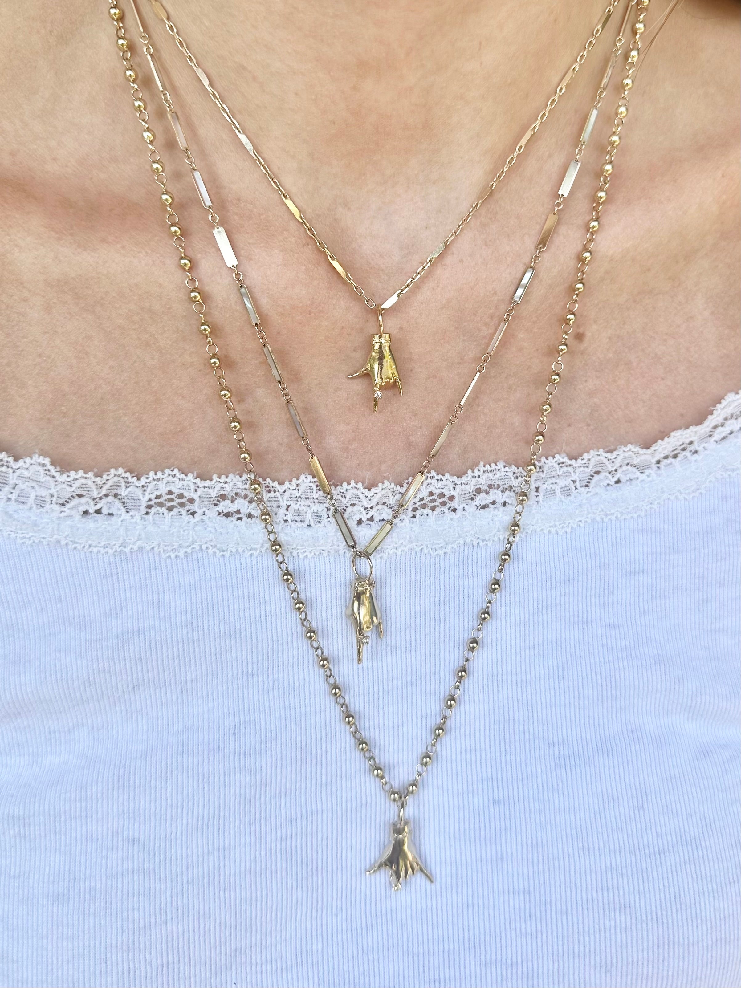 A close-up of a person wearing layered gold necklaces over a white lace-trim top. The necklaces include a beaded chain, a chain with flat rectangular links, and two chain with small bird pendants. Designed by Maura Green