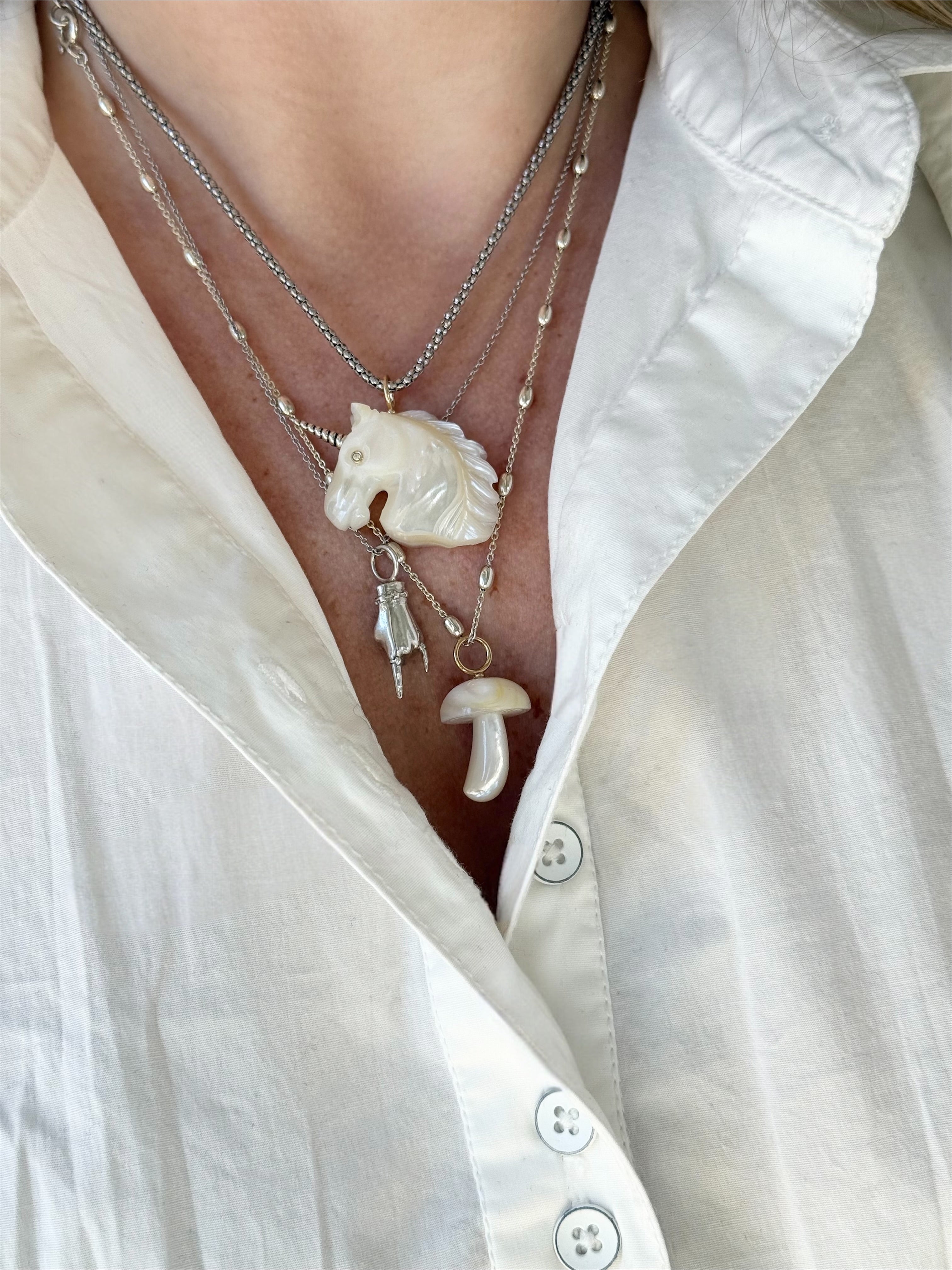 Layered necklaces with white unicorn pendant, silver figural hand pendant making a "rock on" gesture, and a white mushroom pendant, worn by a person in a white shirt. All necklaces designed by Maura Green Jewelry