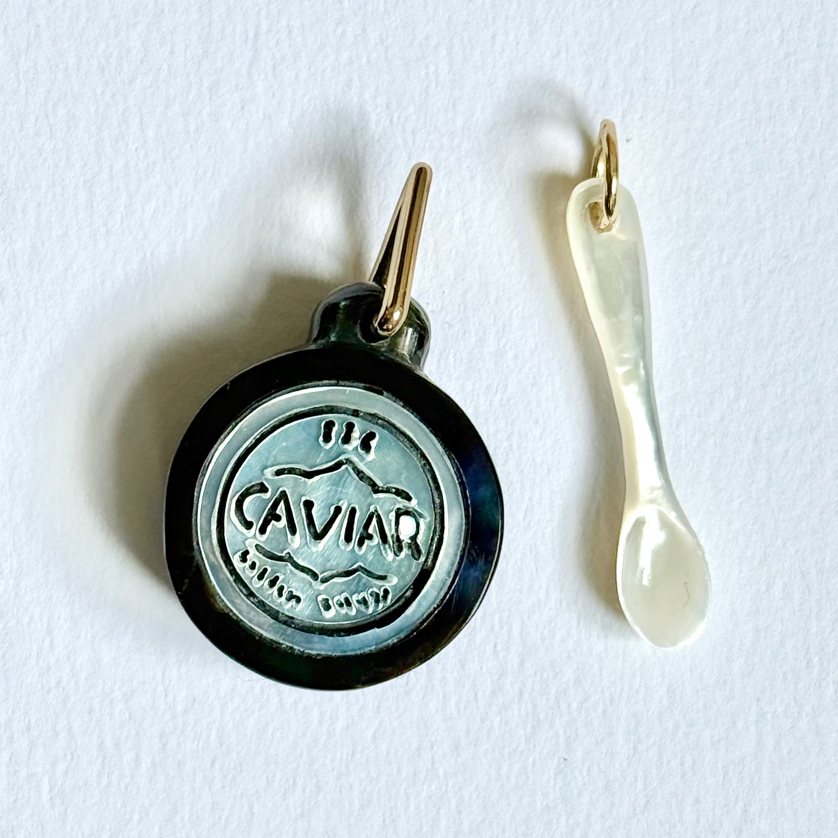 A white mother of pearl caviar spoon charm and a round black and silver-toned caviar tin charm, displayed on a clean white background. Designed by Maura Green Jewelry.